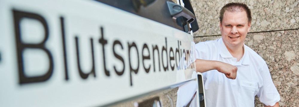 Mann in weißem Shirt steht neben Lastkraftwagen mit Aufschrift ‚Blutspendedienst‘